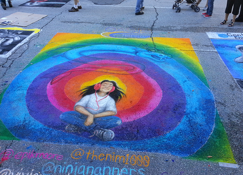 image drawn by a team of artists of a young girl sitting Indian-style listening to music with rainbow colored sound waves behind her at the Via Colori street painting festival 2014 Houston, TX